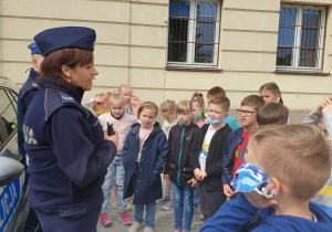 Dzieci ustawione na wybiegu szkolnym słuchając prelekcji policjantki na temat bezpieczeństwa