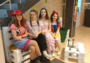 4 dziewczynki ubrane w stroje bajkowe siedzą na ławkach w korytarzu szkolnym