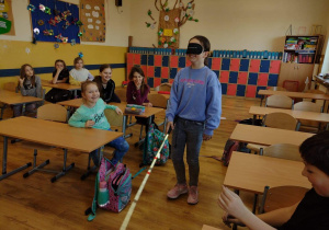 Dziewczynka z zasłoniętymi oczami i laską dla osób niewidomych porusza się po klasie