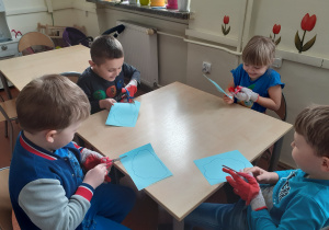 Dzieci siedzą przy stoliku i wycinają niebieskie chmurki z papieru. Na dłoniach założone mają rękawice