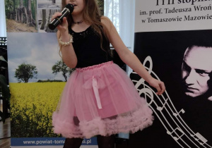 Dziewczynka w kolorowej spódniczce z mikrofonem śpiewa przygotowany utwór