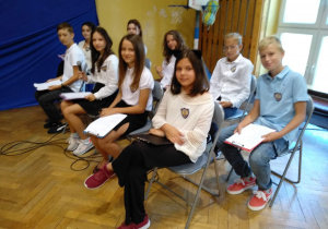Siodmoklasisci siedzą na krzesłach trzymając w dłoniach egzemplarze dzieł Adama Mickiewicza