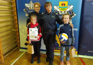 Chłopiec i dziewczynka stojąc z policjantkami prezentują swoje nagrody zdobyte za udział w konkursie
