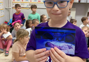 Chłopiec w fioletowej podkoszulce ze swoim zdjęciem w dłoniach jako wcześniak w inkubatorze