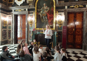 Uczniowie w Zamku Królewskim słuchają pani przewodnik. W tle zabytkowe obrazy z czasów ostatniego króla Polski Stanisława Augusta.