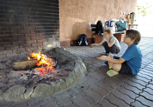 Dziewczynka i chłopiec siedzą przy ognisku i pieką kiełbaski
