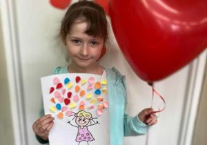 Dziewczynka trzyma w dłoni balon w kształcie serca i pracę plastyczną
